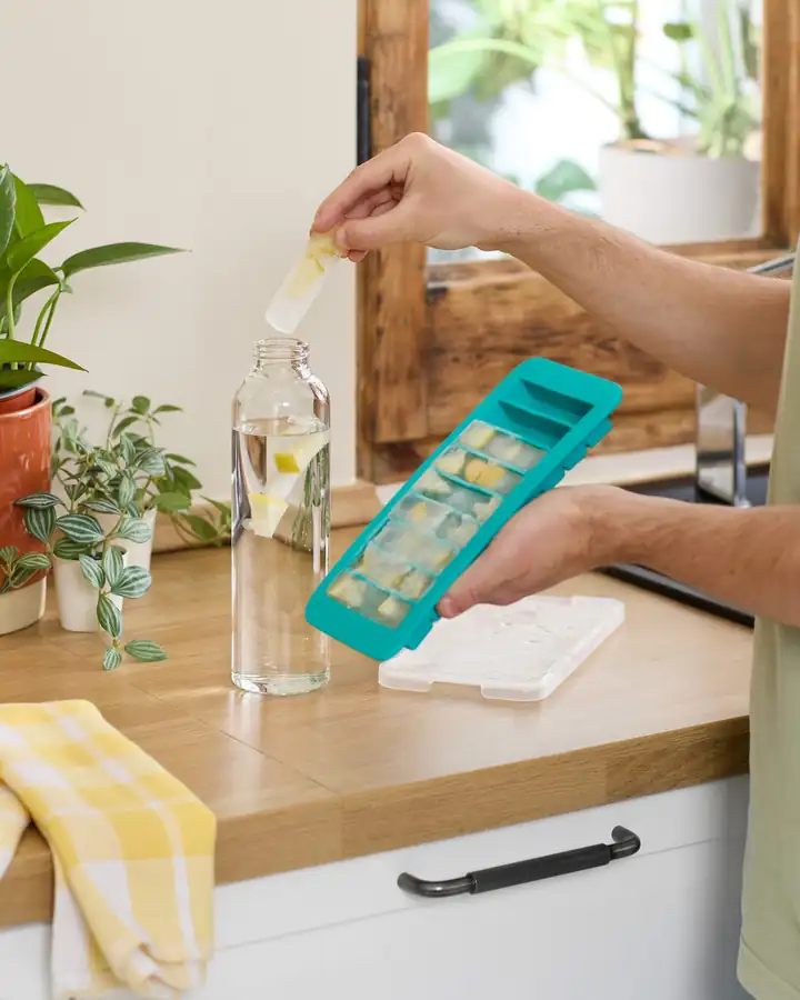 Eiswürfelsticks mit Zitrone aus der türkisen Lékué Form, daneben der Deckel und eine Glasflasche