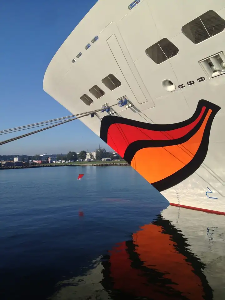 Bug eines AIDA Schiffs mit rotem und orangem Kussmund, der sich im ruhigen Hafenwasser spiegelt