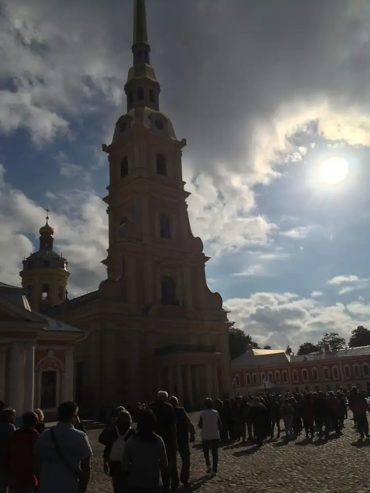 Peter und Paul Kathedrale in St. Petersburg mit goldener Turmspitze vor dunklen Wolken, davor wartende Besucher