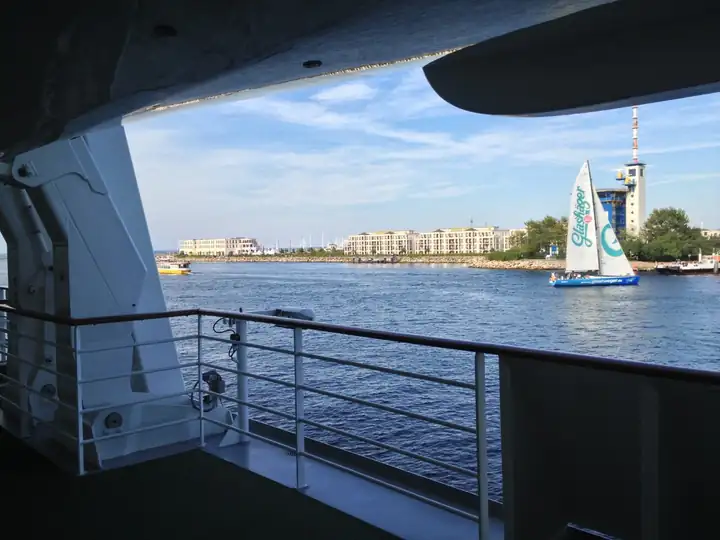 Ausfahrt aus Warnemünde vom Deck eines Kreuzfahrtschiffs, daneben ein Segelboot und ein Turm an der Hafeneinfahrt