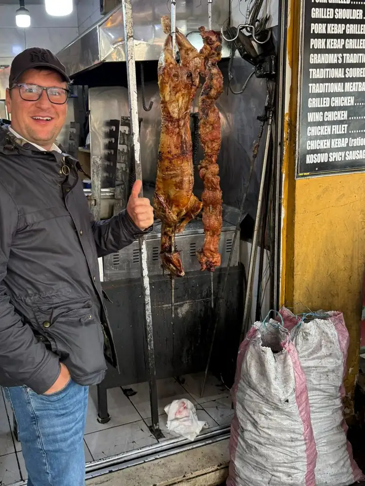 Lars Laschinski zeigt mit dem Daumen nach oben neben Lamm am Spieß in einer Grillstube, rechts eine Tafel mit Gerichten vom Grill