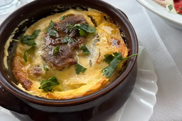 Überbackenes Gericht mit Petersilie in einem braunen Tontopf, daneben eine Schüssel Salat mit Tomaten und Zwiebeln