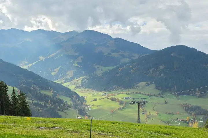 Blick von einer Almwiese über ein grünes Tal mit Wald und Wiesen, dahinter Bergketten unter Wolken, rechts ein Liftmast