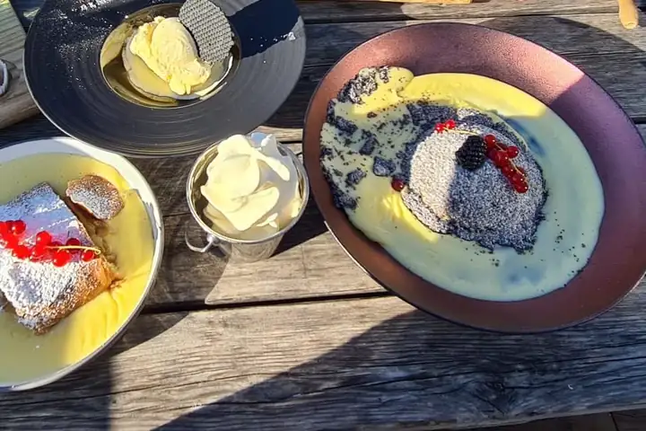 Holztisch in der Sonne mit Apfelstrudel in Vanillesoße mit roten Johannisbeeren, einem Germknödel mit Mohn in Vanillesoße und Vanilleeis in einem Becher