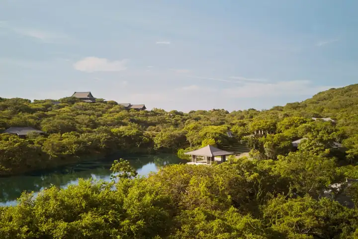 Bewaldete Hügel an einem See, dazwischen ein offener Pavillon und mehrere Dächer im Grün