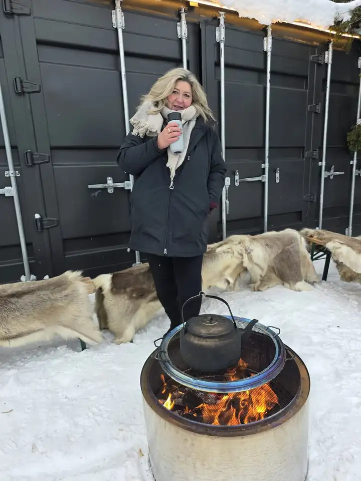 Anja Kosmol im Schnee an einer Feuerschale mit Wasserkessel in Åre, hinter ihr Bänke mit Fellen