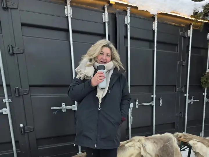 Anja Kosmol im Schnee mit einem heißen Becher in der Hand, hinter ihr dunkle Containerwände und Bänke mit Fellen