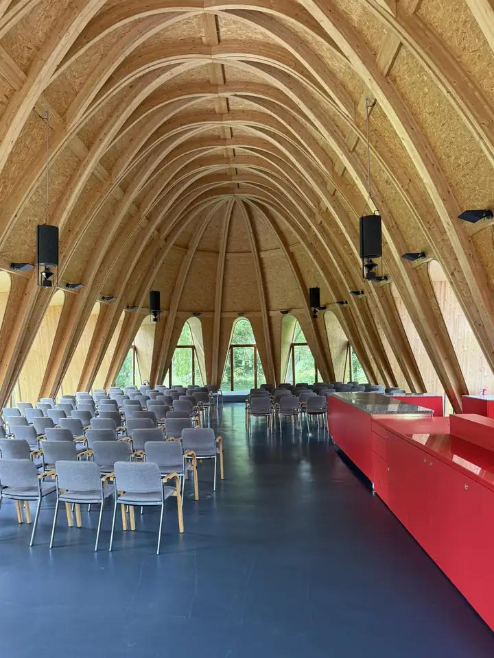 Hoher Saal mit gebogenen Holzträgern, Stuhlreihen auf dunklem Boden, rechts eine rote Theke, hinten hohe Fenster ins Grüne