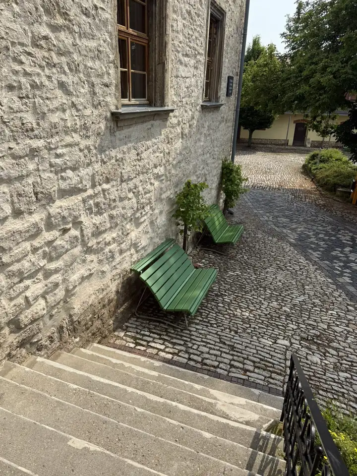 Zwei grüne Holzbänke an einer hellen Bruchsteinmauer, davor eine Treppe und ein gepflasterter Weg