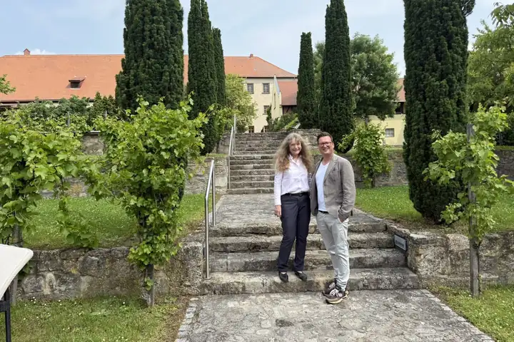 Marion Schneider und Lars Laschinski vor einer Steintreppe im Garten, umgeben von Säulenzypressen und Reben, dahinter das Schloss