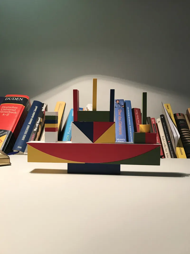 Bauhaus Bauspiel als Schiff aufgebaut: roter Bogen als Rumpf auf einem flachen Sockel, darauf Dreiecke in Blau, Gelb und Rot, Quader und dünne Stäbe als Masten, dahinter ein Bücherregal