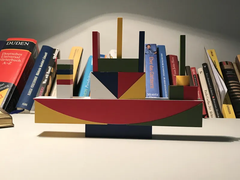 Bauhaus Bauspiel als Schiff aufgebaut vor einem Bücherregal: roter Bogen, Dreiecke, Quader und Stäbe in Rot, Gelb, Blau und Grün