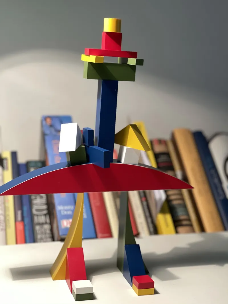 Bauhaus Bauspiel als Turm: ein blauer Stab auf einem roten Bogen, oben grüne und rote Teile und ein gelber Zylinder, unten Füße aus kleinen Quadern