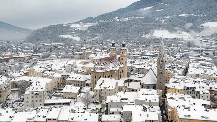 Brixen im Schnee von oben, in der Mitte die Türme von Dom und Pfarrkirche, dahinter verschneite Berge