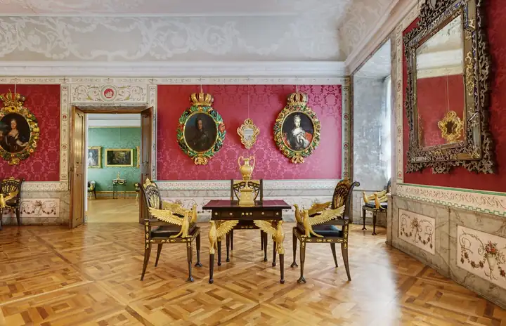 Raum im Kaisertrakt der Hofburg Brixen mit roter Wandbespannung, Porträts in Goldrahmen, Spiegel und Tisch auf Parkett