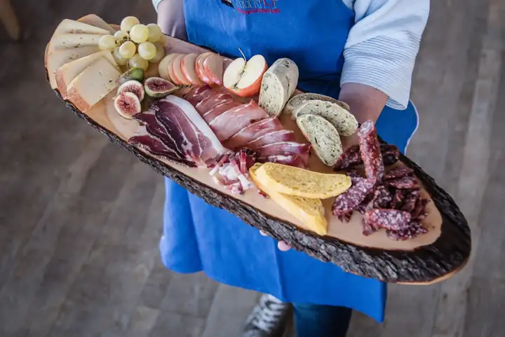Holzbrett mit Speck, Wurst, Käse, Schüttelbrot, Trauben und Feigen, gehalten von einer Person in blauer Schürze