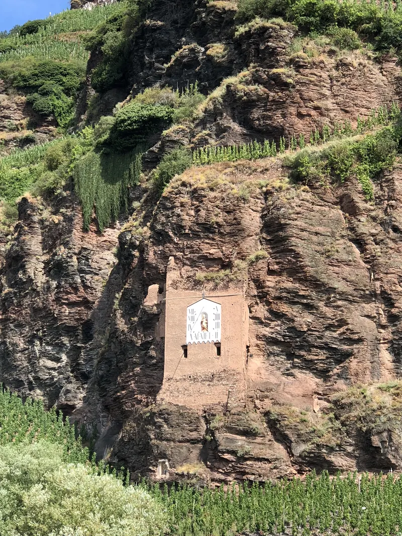 Roter Felsen zwischen Weinbergen mit einem gemauerten Turm, darauf eine weiße Sonnenuhr mit einer Figur in einer Nische