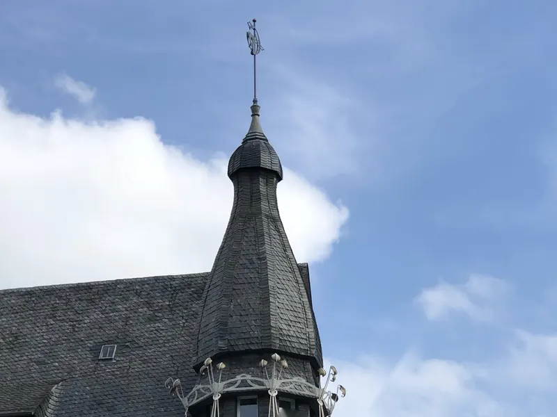 Spitzer Turm aus Schiefer mit Wetterfahne vor blauem Himmel, darunter ein Kranz aus geschwungenen Eisenstäben
