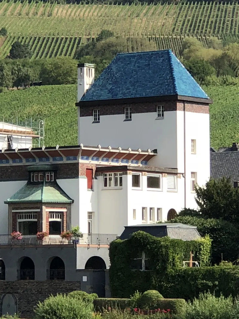Weiße Villa mit blauem Walmdach und Erker vor steilen Weinbergen, davor Gärten und eine bewachsene Mauer