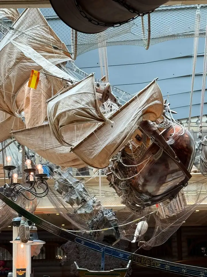 Großes Segelschiff mit hellen Segeln und Netzen unter einer Glasdecke, unten eine Laterne mit dem Schriftzug Bell Rock