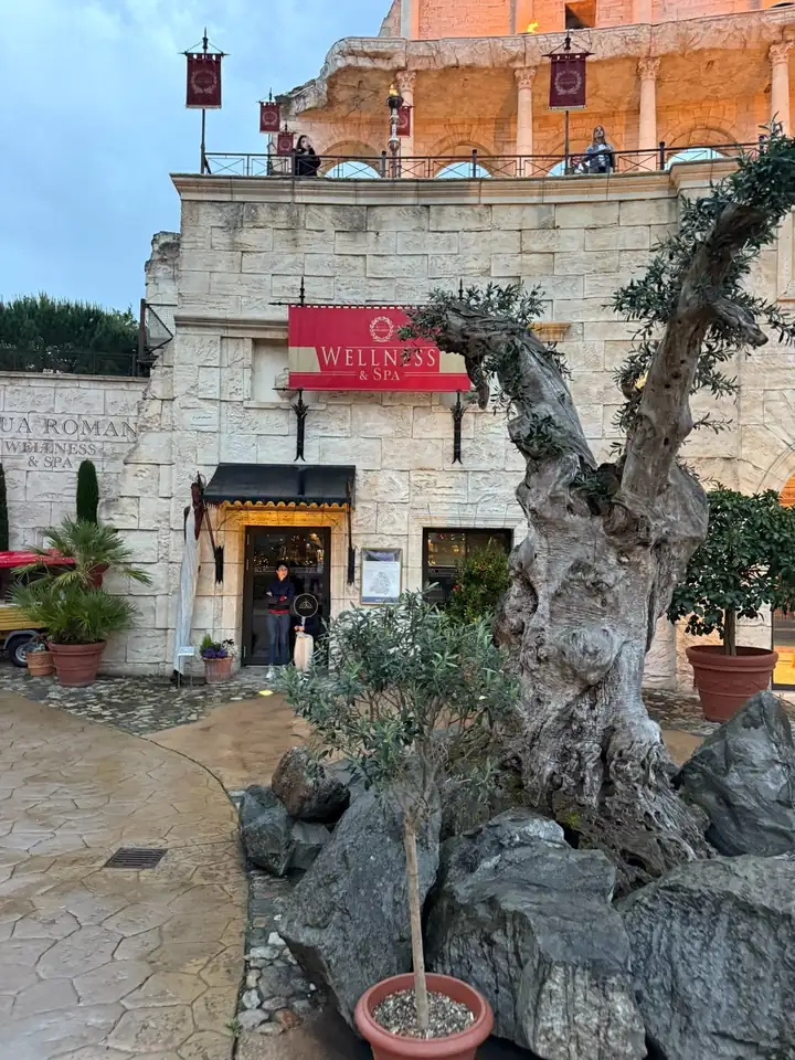 Eingang zum Wellnessbereich Aqua Romana im Hotel Colosseo mit Mauern im römischen Stil, einem roten Schild und einem alten Olivenbaum