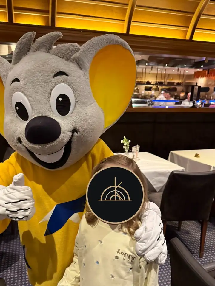 Ein Kind neben dem Maskottchen Ed Euromaus im Restaurant, das Gesicht des Kindes ist mit dem GRAND KIN Logo verdeckt