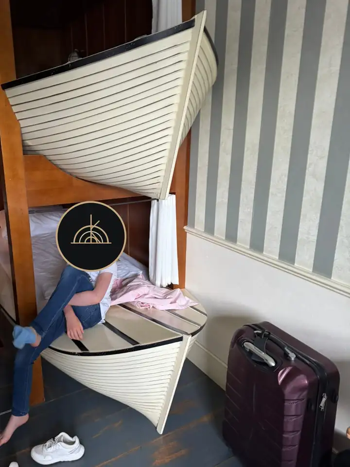 Zwei Betten in Form weißer Ruderboote übereinander in einem Hotelzimmer mit gestreifter Tapete, im unteren Boot sitzt ein Kind, sein Gesicht ist mit dem GRAND KIN Logo verdeckt, rechts ein Koffer