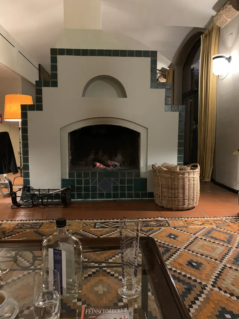Offener Kamin mit brennendem Feuer in einer weißen Wand mit grünen Fliesen in Stufenform, davor ein Teppich mit Rautenmuster und ein Glastisch