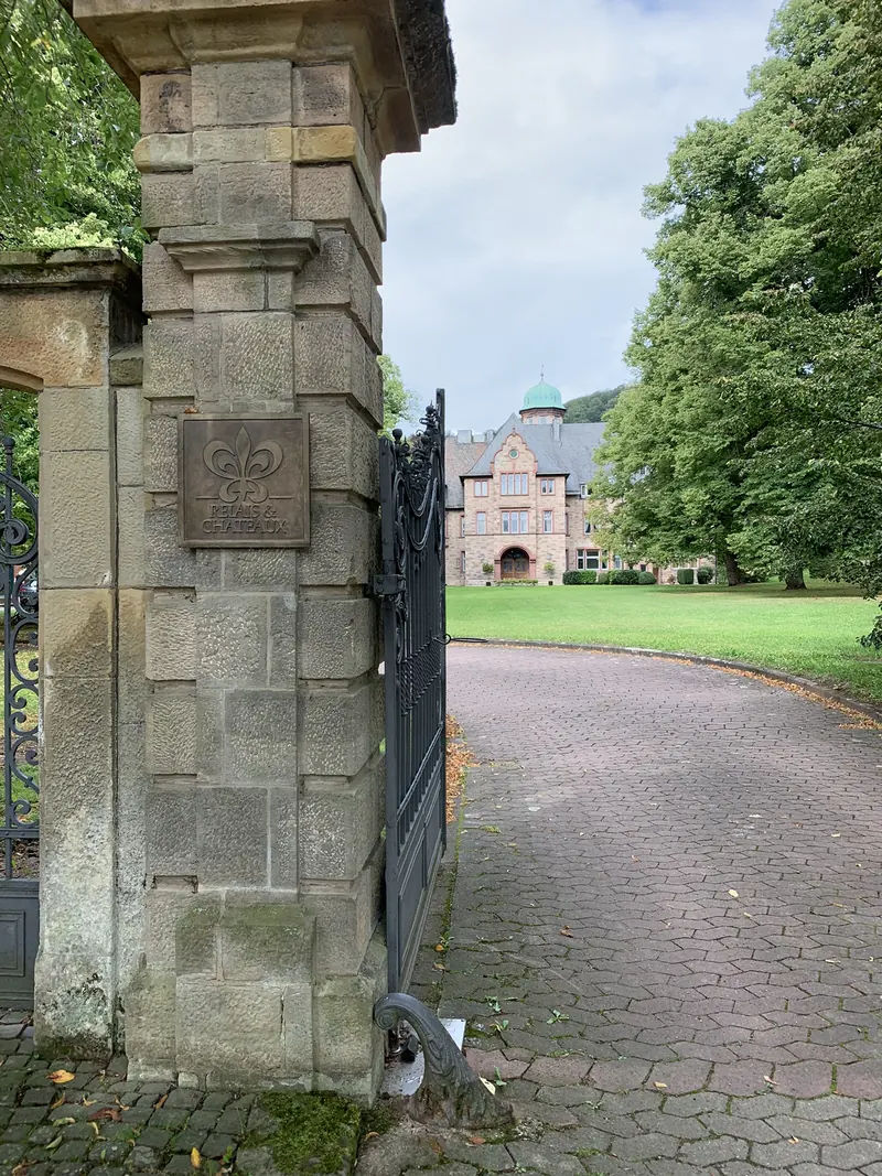 Steinpfeiler mit Bronzeschild und schmiedeeisernem Tor, dahinter eine gepflasterte Auffahrt, Rasen und das Schloss Hohenhaus mit grüner Kuppel