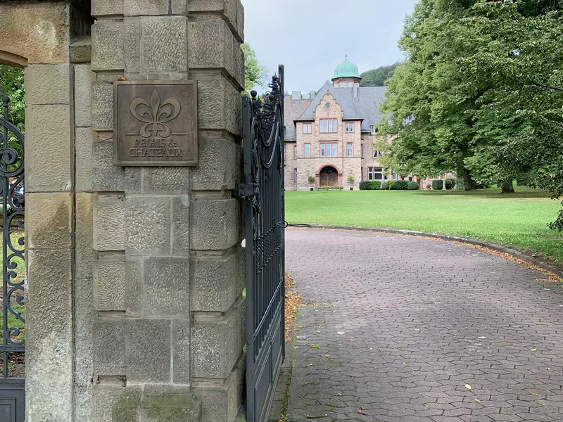 Steinpfeiler mit Bronzeschild am Tor, dahinter die Auffahrt und das Schloss Hohenhaus mit grüner Kuppel