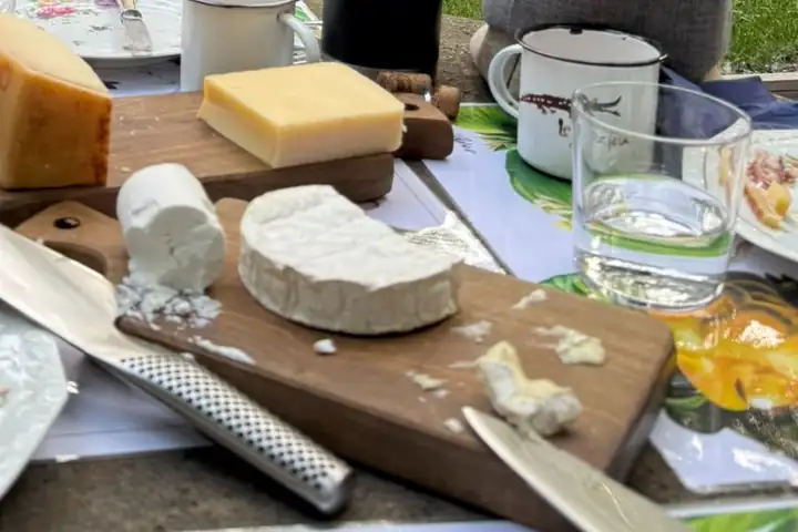 Ein großes Messer mit Griff aus Stahl neben einem Holzbrett mit Ziegenkäse und Weichkäse, dahinter ein Block Schnittkäse, ein Becher aus Emaille und ein Wasserglas