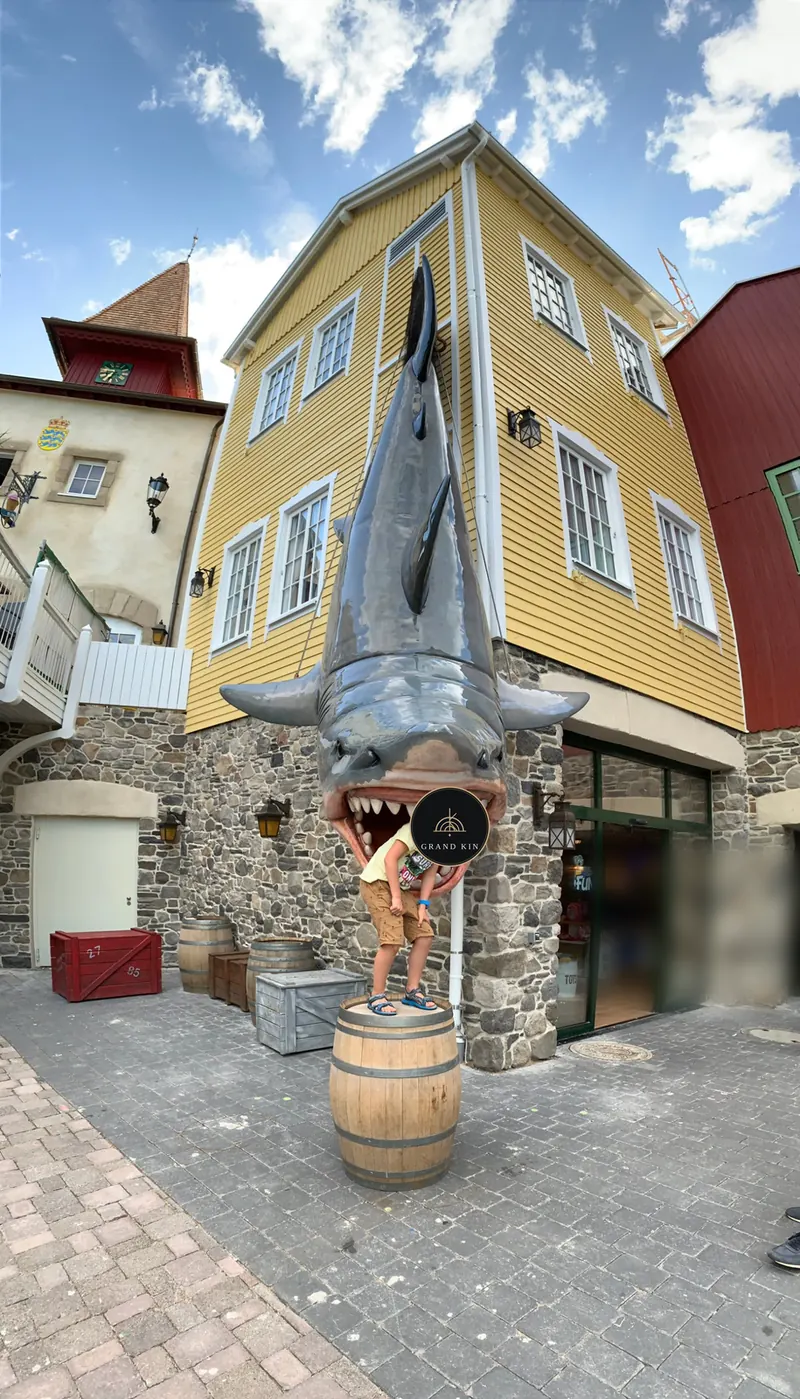 Großer Hai als Figur über dem Eingang eines gelben Holzhauses, ein Kind steht auf einem Fass im Maul des Hais, das Gesicht ist mit einem Logo verdeckt