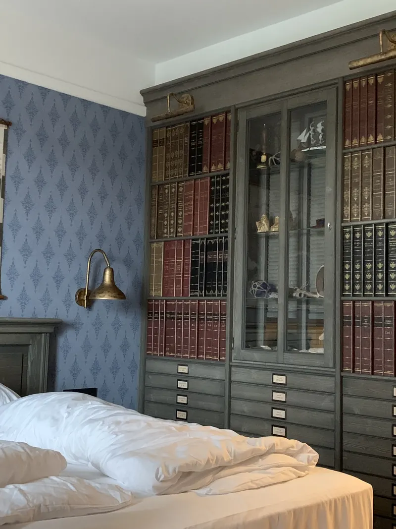Hotelzimmer mit blauer Mustertapete und einem großen Schrank mit Büchern, Vitrinen und Schubladen