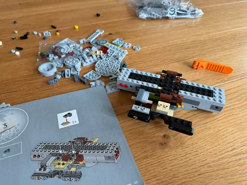 Halb gebauter Kern des Millennium Falcon aus grauen, schwarzen und braunen Steinen neben dem aufgeschlagenen Bauheft und einem orangen Steinetrenner