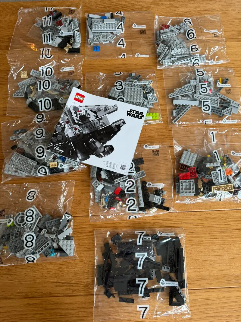 Elf nummerierte Plastiktüten mit grauen, schwarzen und bunten LEGO Steinen auf einem Holztisch, in der Mitte das Bauheft mit dem Millennium Falcon