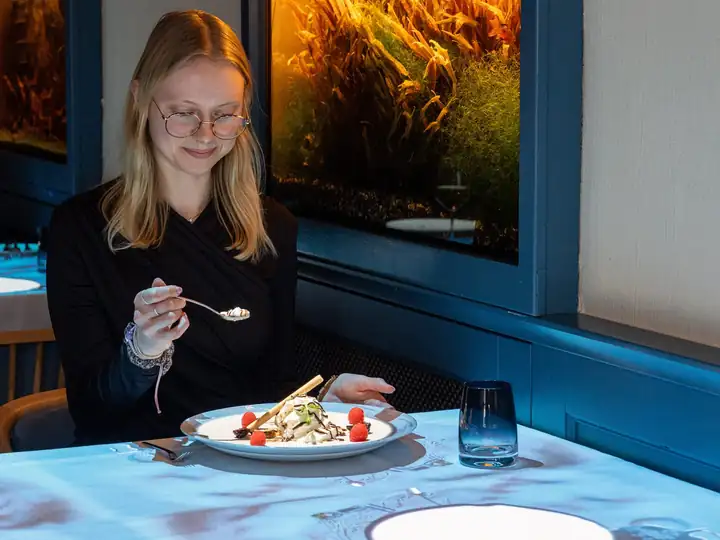 Junge Frau mit Brille isst ein Dessert mit Vanilleeis und Himbeeren an einem Tisch mit blau leuchtender Projektion, dahinter ein beleuchtetes Aquarium