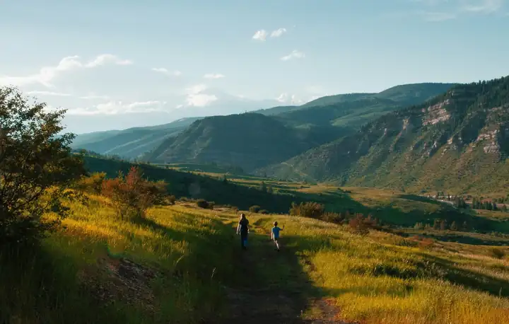 Mutter und Sohn wandern von hinten gesehen auf einem Wiesenpfad durch ein Bergtal im Abendlicht