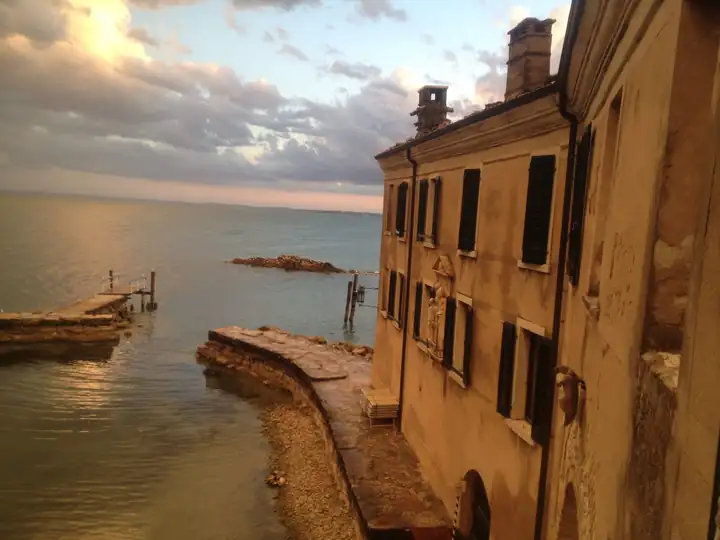 Punta San Vigilio am Abend: Steinmole und Steg im goldenen Licht, dahinter der Gardasee unter Wolken, rechts die Locanda