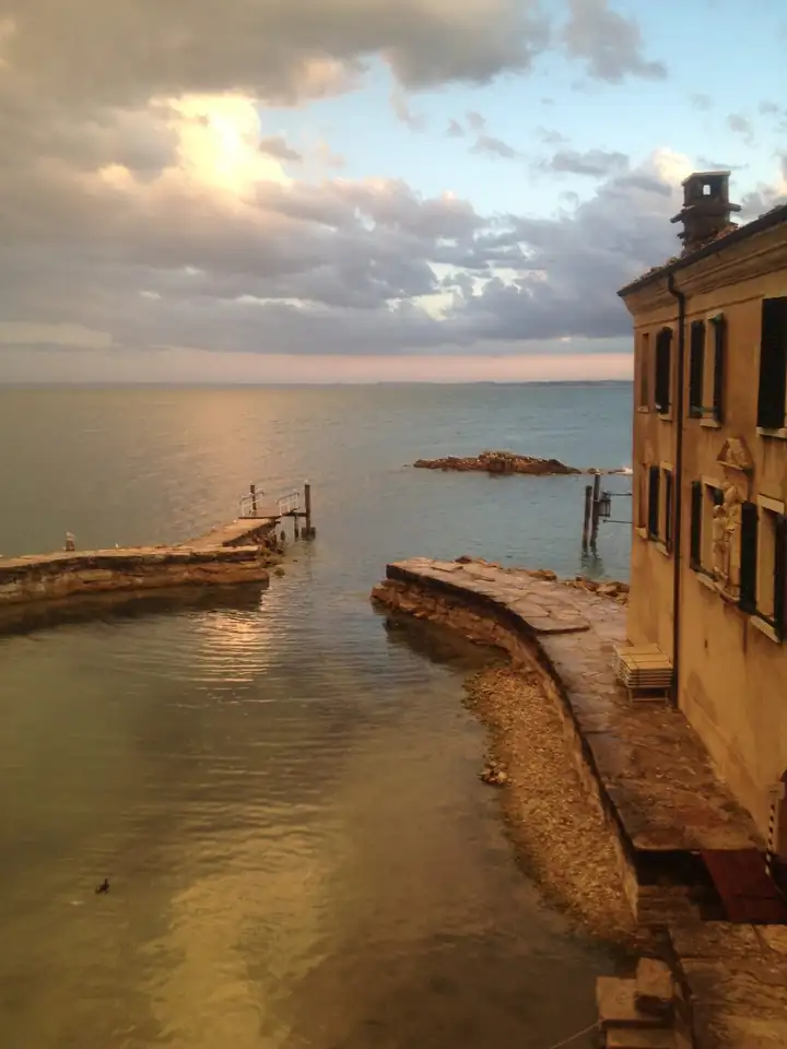 Abendlicht über dem Gardasee, eine geschwungene Steinmole führt am Haus entlang ins Wasser, rechts die Fassade der Locanda