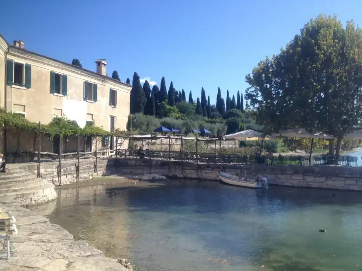 Kleiner Hafen von Punta San Vigilio mit hellem Steinbecken, einem Holzboot, dahinter die Locanda mit grünen Fensterläden, Zypressen und Platanen