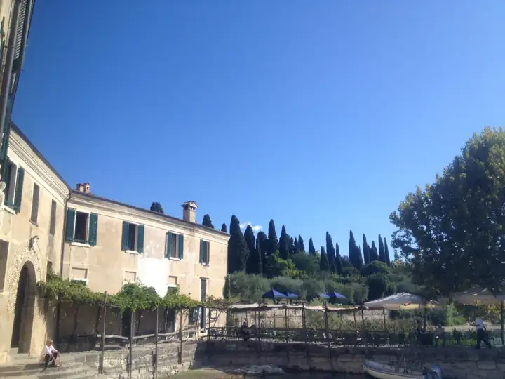 Fassade der Locanda San Vigilio mit grünen Fensterläden und Torbogen, davor die Terrasse mit Sonnenschirmen und Zypressen