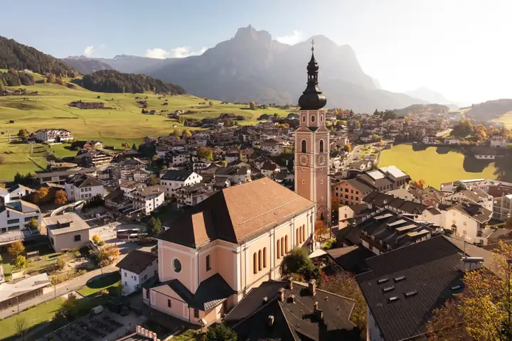 Blick von oben auf Kastelruth mit rosafarbener Kirche und hohem Kirchturm, dahinter grüne Wiesen, Wälder und die Dolomiten