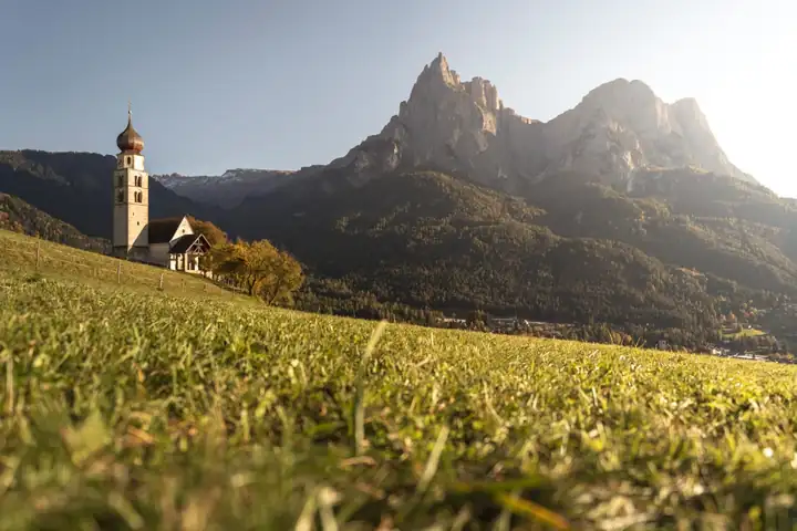 Das Kirchlein St. Valentin mit Zwiebelturm auf einer Wiese oberhalb von Seis, dahinter die Felsen des Schlern im Gegenlicht
