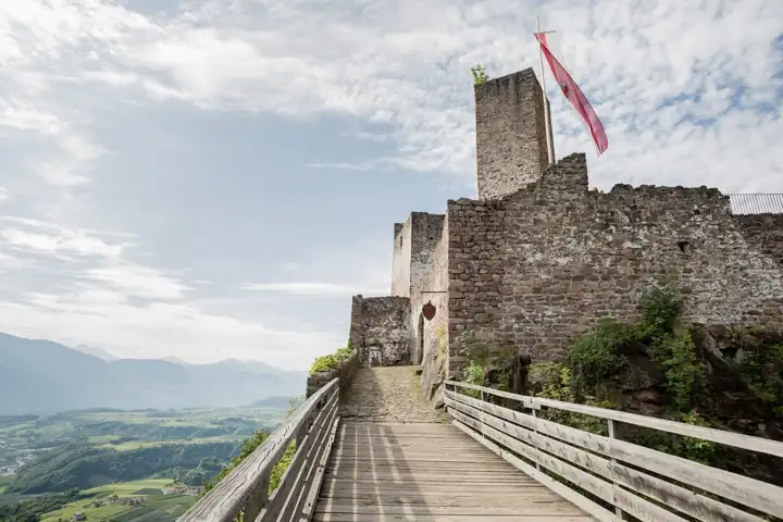 Holzsteg entlang der Mauern von Burg Hocheppan mit Bergfried und roter Fahne, links der Blick ins Tal