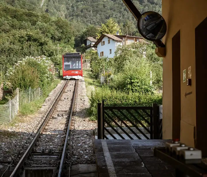 Roter Wagen der Standseilbahn auf die Mendel fährt auf dem Gleis zwischen Gärten auf die Station zu
