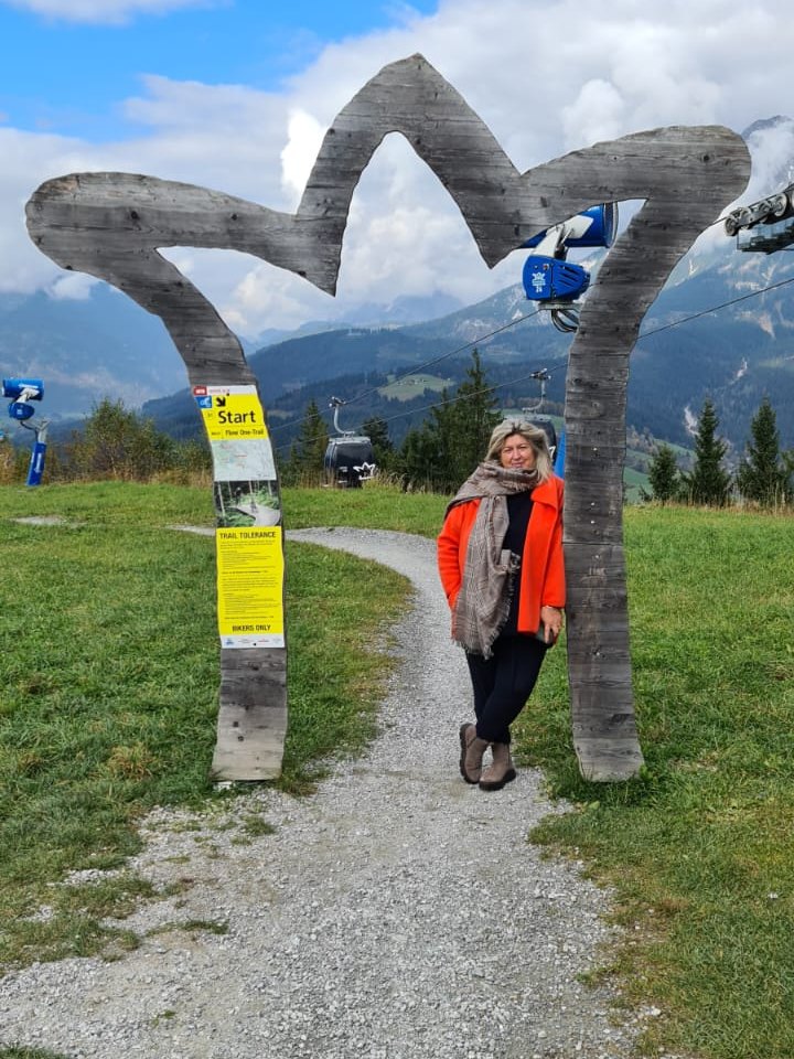 Anja Kosmol in orangefarbener Jacke an einem großen Holzbogen in Form einer Krone auf einer Almwiese, dahinter Berge und eine Gondel