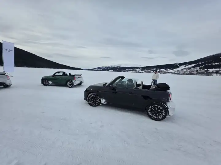 Ein schwarzes und ein grünes MINI Cabrio mit offenem Verdeck auf dem verschneiten See, links eine Fahne mit MINI Logo, hinten Berge