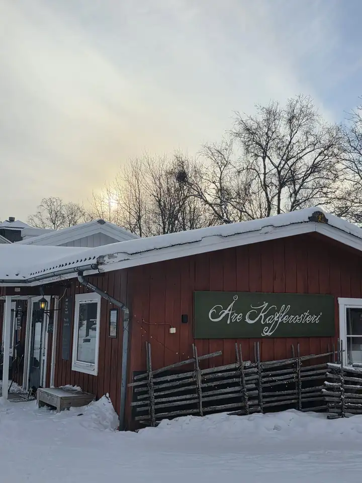 Rotes Holzhaus der Åre Kafferosteri mit grünem Schild, davor Schnee und ein Zaun aus Ästen, dahinter kahle Birken im Gegenlicht