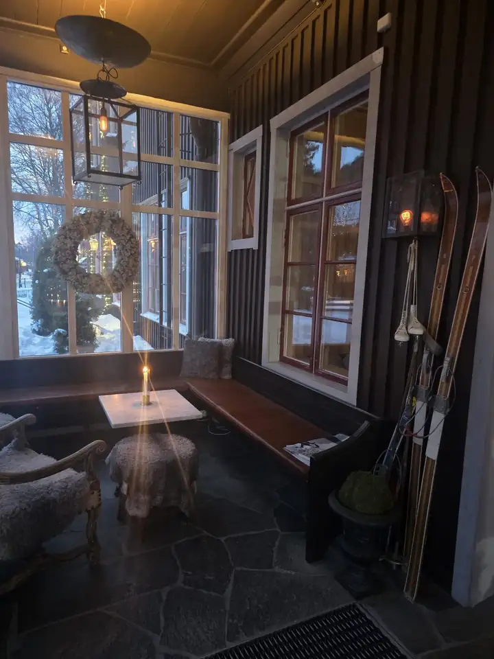 Verglaste Veranda mit Kerze auf einem Marmortisch, Fellhocker, Holzbank, einem Kranz im Fenster und alten Holzski an der dunklen Holzwand