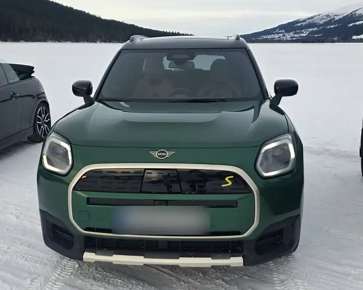 Grüner MINI Countryman frontal auf dem verschneiten Eis, dahinter Wald und Berge mit Schnee